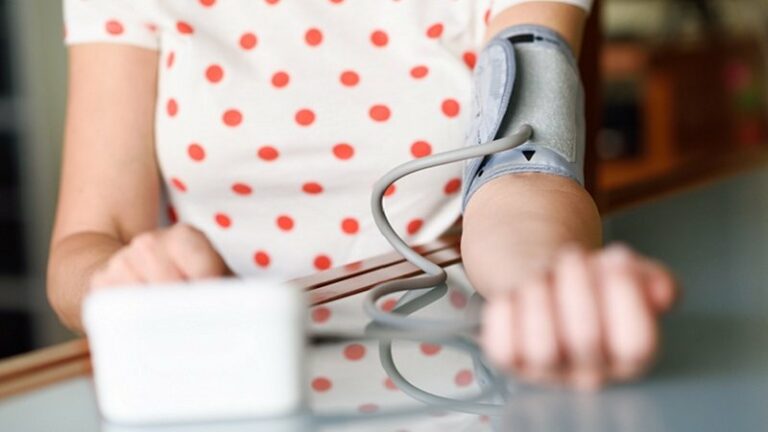 Bluthochdruck_web_800.jpg Eine Person mit einem Blutdruckmessgerät am Arm, das auf einem Tisch liegt. Im Hintergrund ein unscharfer Raum.