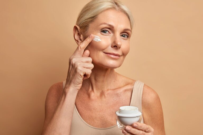 Anti-Aging.jpg Eine Frau mit blondem Haar trägt Gesichtspflegecreme auf ihre Wange auf, während sie einen Cremetiegel in der Hand hält.