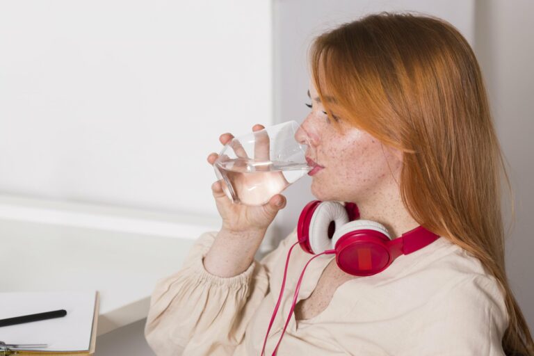 fluessigkeitsmangel.jpg Eine Person mit roten Kopfhörern trinkt aus einem Glas Wasser und sitzt an einem Schreibtisch.