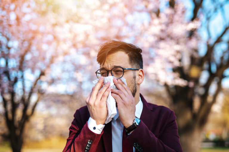 Heuschnupfen.jpg Ein Mann in einem Anzug benutzt ein Taschentuch, um sich die Nase zu putzen, vor blühenden Bäumen im Hintergrund.