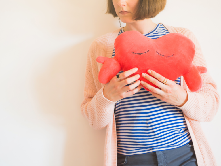 Eine Person hält ein rotes herzförmiges Kissen mit geschlossenen Augen, gekleidet in gestreiftes Oberteil und eine helle Strickjacke.